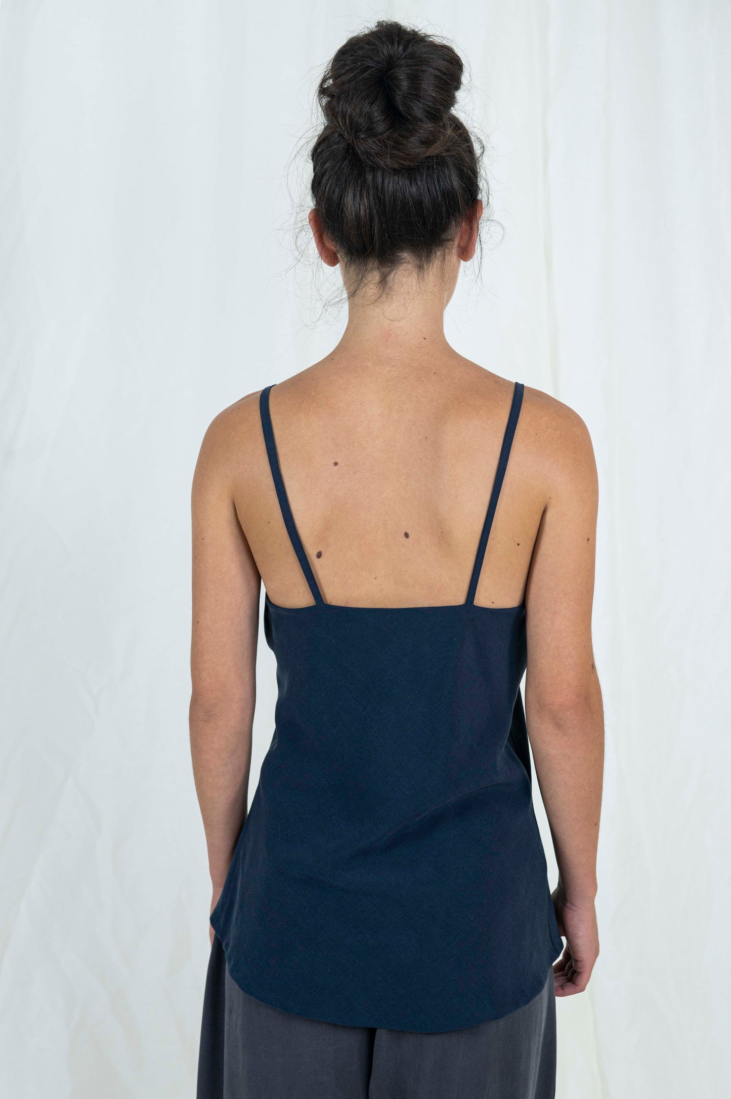 Close up back view of woman wearing bias cut camisole tank top in dark indigo tencel fabric. Moonstone grey tencel pants can be seen.