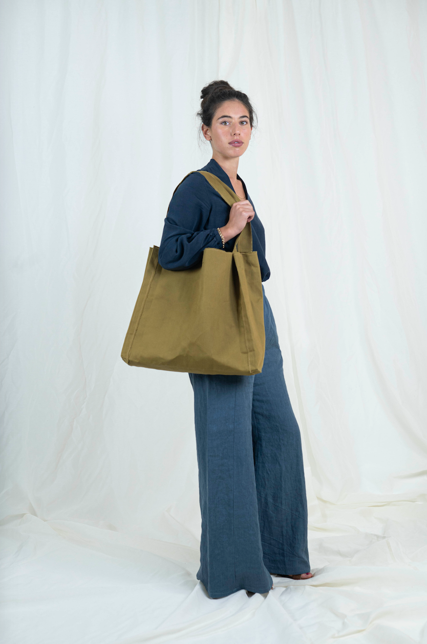 Dark haired woman wearing medium blue wide legged linen pants, dark blue bishop sleeve top and oversized tan canvas tote bag, viewed from the side, gaze is forward.