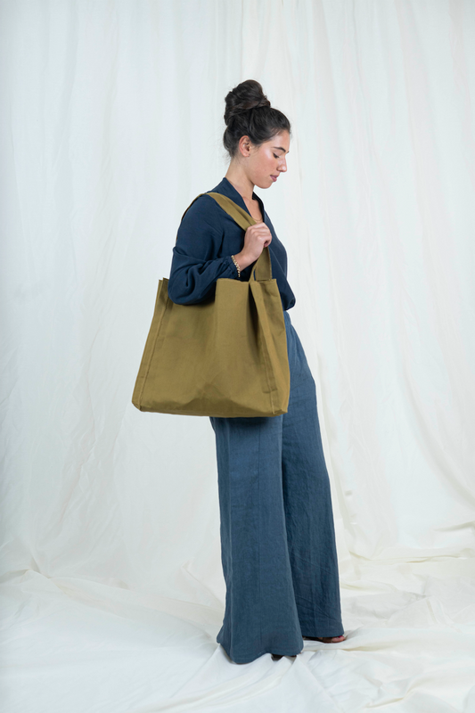Dark haired woman wearing medium blue wide legged linen pants, dark blue bishop sleeve top and oversized tan canvas tote bag, viewed from the side, gaze is downward.